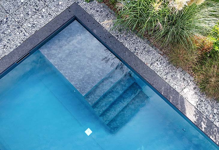 Poolfolie makellos in ein modernes Betonschwimmbecken eingelegt, mit klarem Wasser, flankiert von Kiesbett und üppigen Gräsern vor einer Terrassen-Lounge.