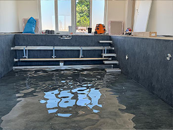 Innenbereich einer Schwimmhalle mit neu verlegter, anthrazitgrauer Poolfolie an Wänden und Boden, teilweise gefüllt mit Wasser, im Hintergrund Gegestell für Treppe und Rollladenschacht.