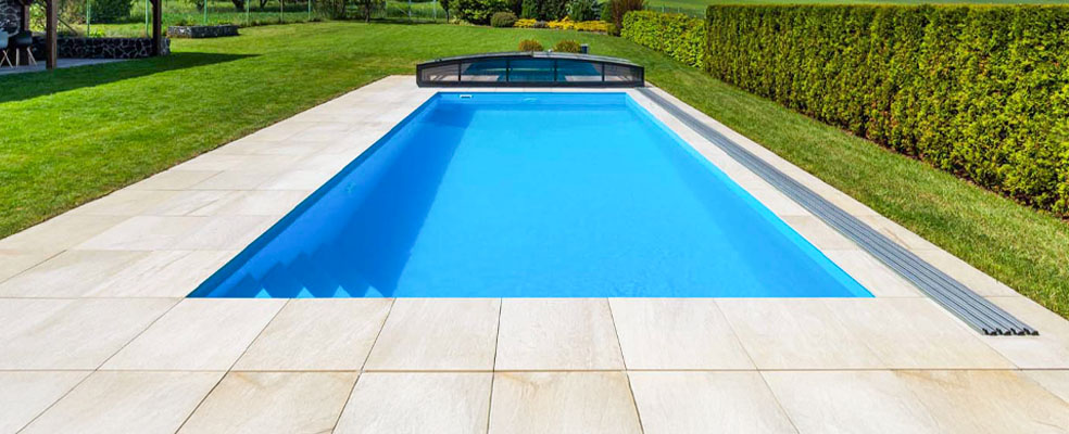 PP Pool Albixon: eingelassener, rechteckiger Polypropylen-Pool mit kristallklarem, türkisfarbenem Wasser, hellen Natursteinplatten und teilgeöffneter, dunkelgrauer Schiebeabdeckung vor grünem Rasen und Thuja-Hecke.