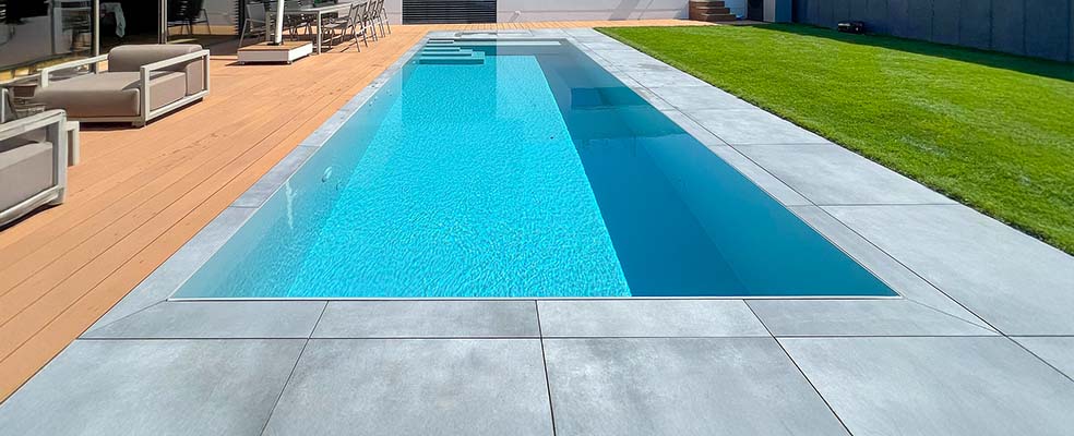 PP Pool Elite: moderner, eingelassener Polypropylen-Pool mit glasklarem Wasser, integrierter Sitz- und Liegefläche, anthrazitgrauer Plattenumrandung und angrenzender Holzterrasse vor zeitgenössischer Hausfassade.