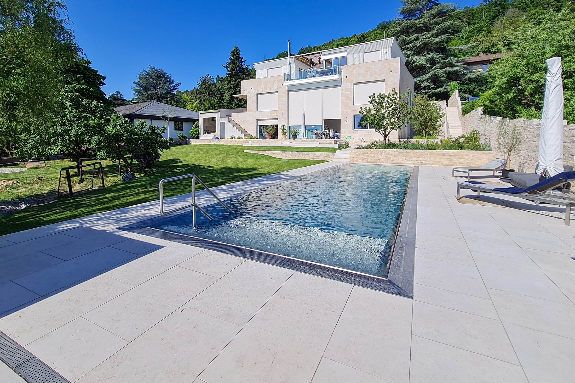 Edelstahlpool mit Luftsprudelliege, gerader Treppe und Blick auf eine moderne Villa mit Gartenanlage