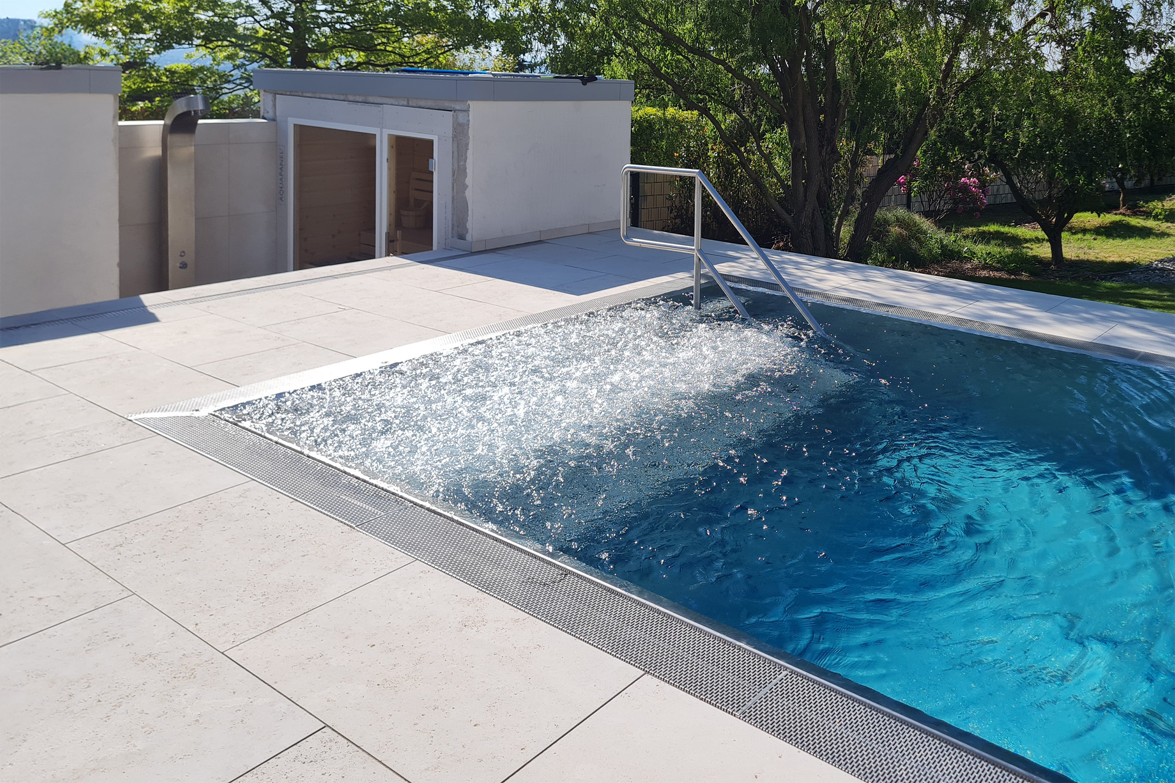 Edelstahl-Überlaufbecken mit eingeschalteter Luftsprudelliege, Treppeneinstieg mit Handlauf und Blick auf die Sauna im Hintergrund