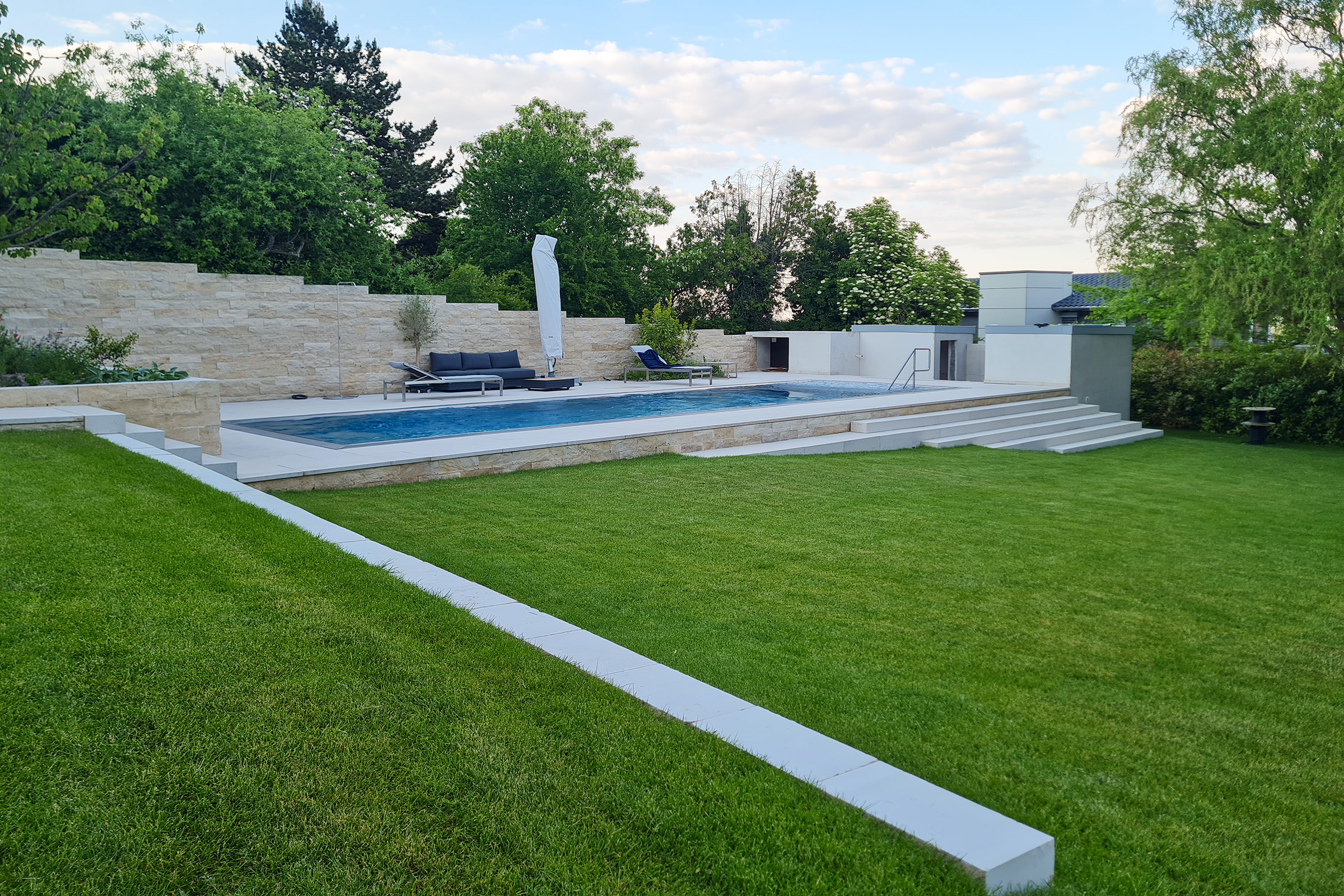 Edelstahlpool als Überlaufbecken mit breitem Einstiegsbereich und großzügigem Gartenumfeld mit Natursteinmauer