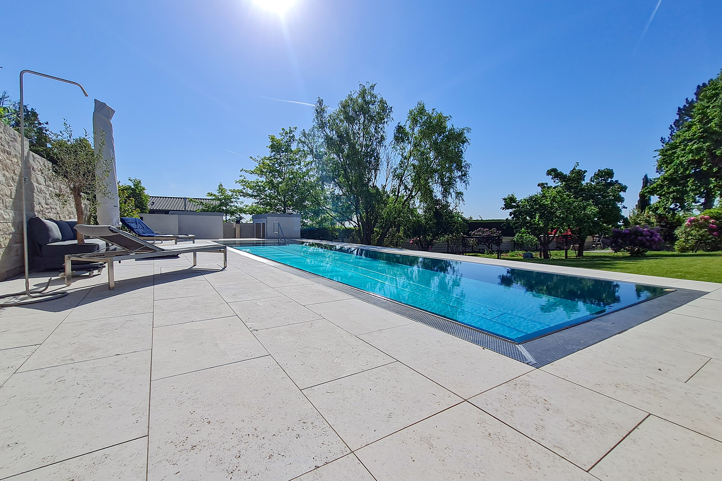 Flacher Edelstahl-Überlaufpool mit reflektierender Rollladenabdeckung in der Nachmittagssonne, Garten und Lounge im Hintergrund