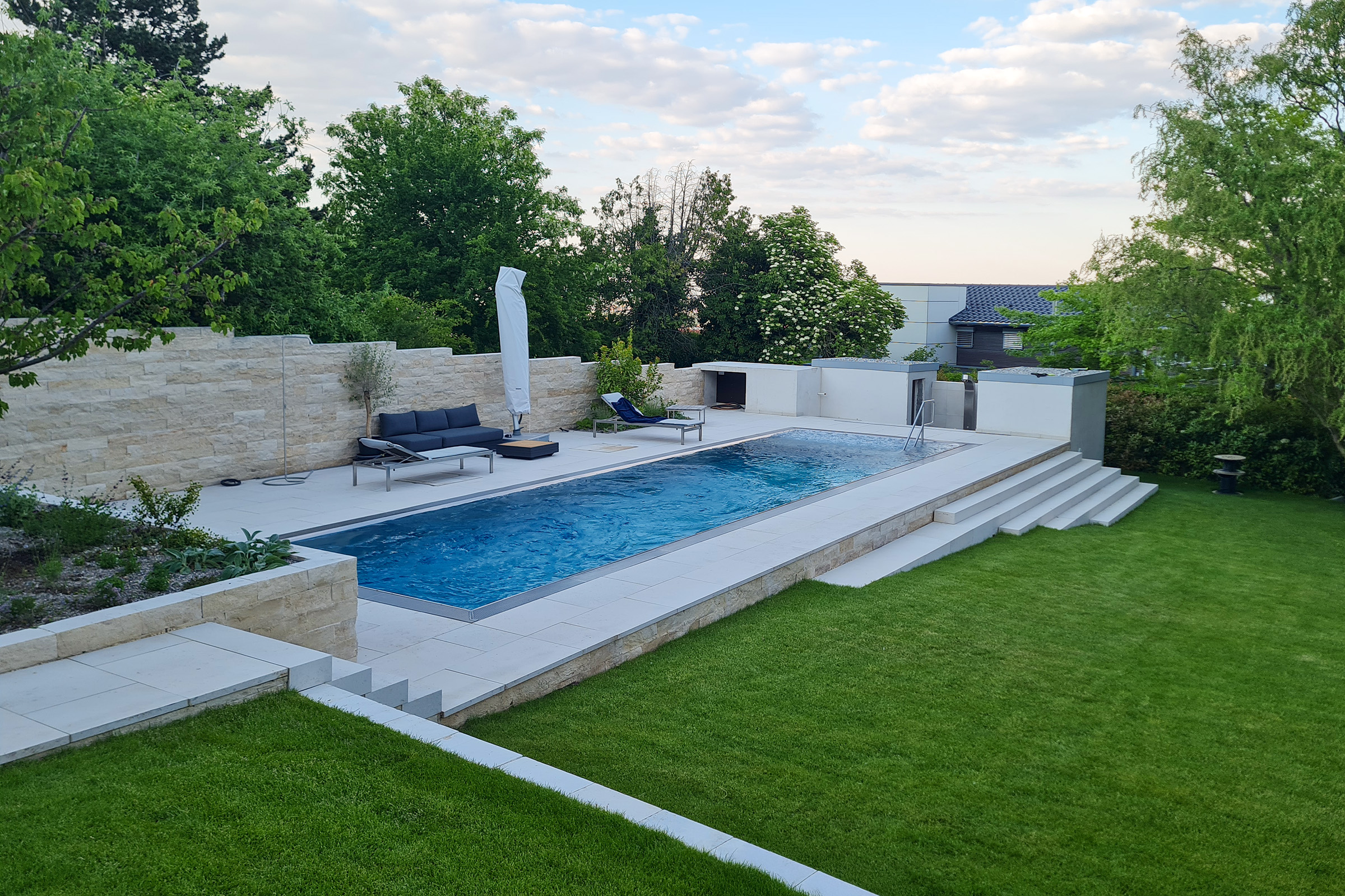 Großzügiges Edelstahlschwimmbecken mit Überlaufrinne und Sitzecke, eingefasst von Natursteinmauern und Gartenterrassen